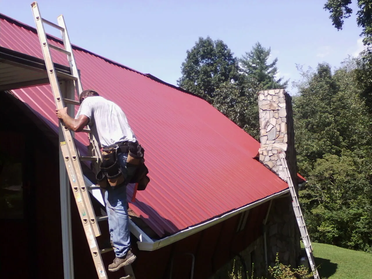 Roofer on a ladder beside a red metal roof and stone chimney — Soffit & Fascia Repair by Roofing Brothers in Due West